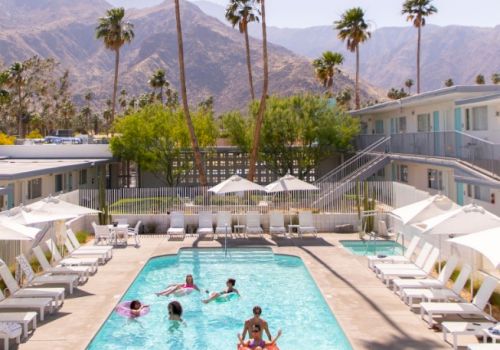 A group of people enjoy a sunny day at a hotel pool surrounded by lounge chairs, with palm trees and mountains in the background.