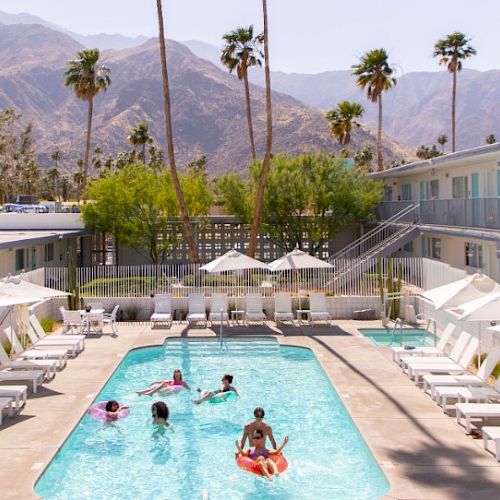 A group of people enjoy a sunny day at a hotel pool surrounded by lounge chairs, with palm trees and mountains in the background.