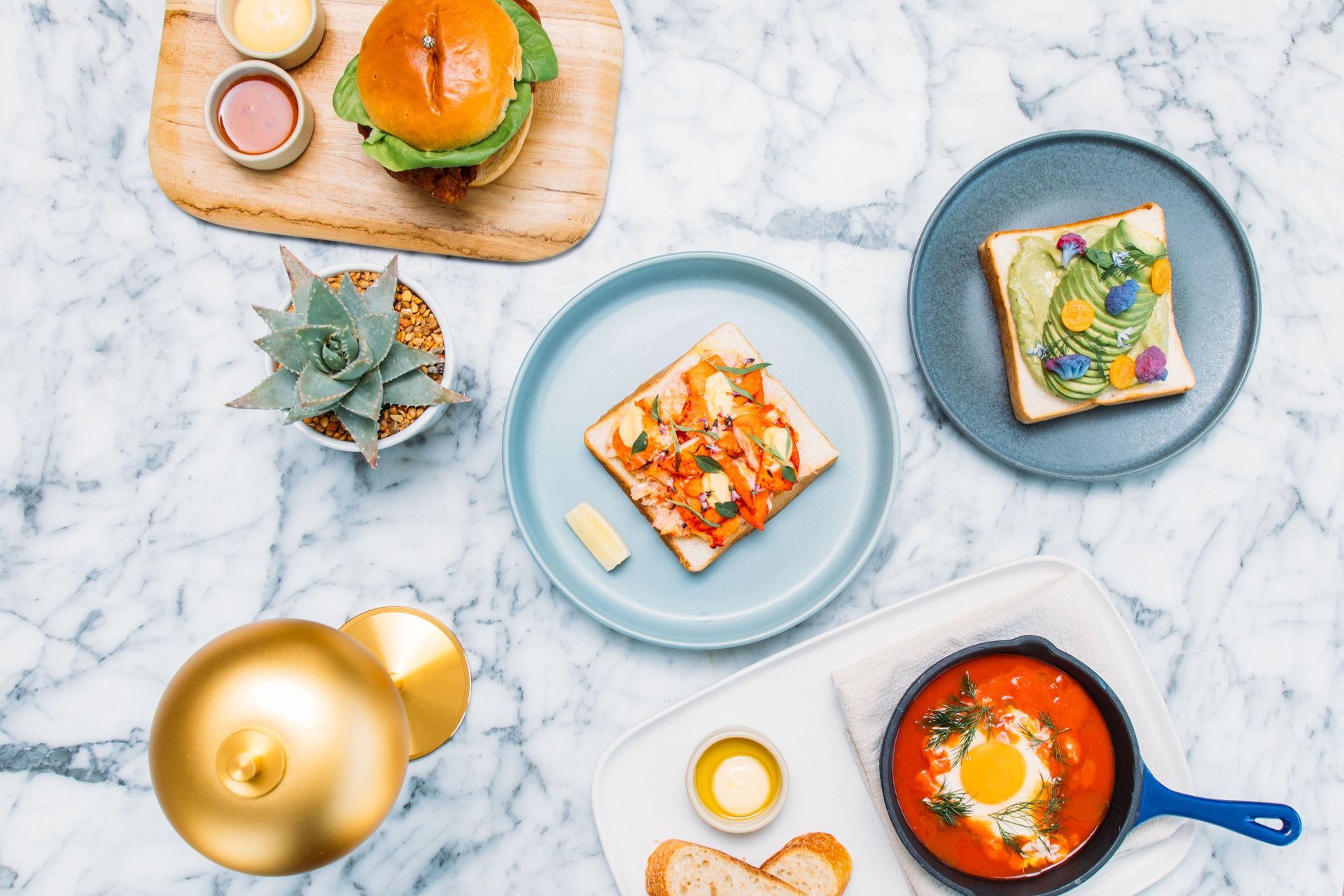 The image shows a variety of dishes including toast with toppings, a burger, a small skillet meal, and a plant on a marble surface.