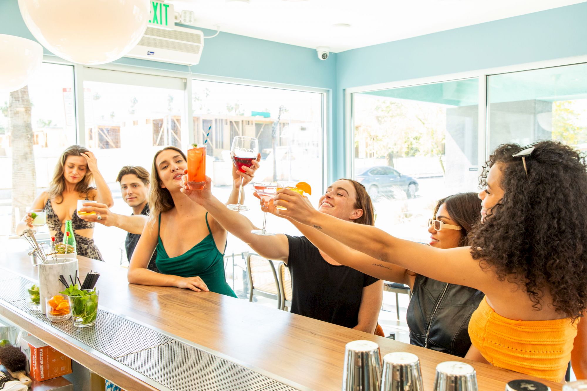 A group of people is gathered at a bar, raising their drinks in a celebratory toast while smiling and socializing in a bright, casual indoor setting.