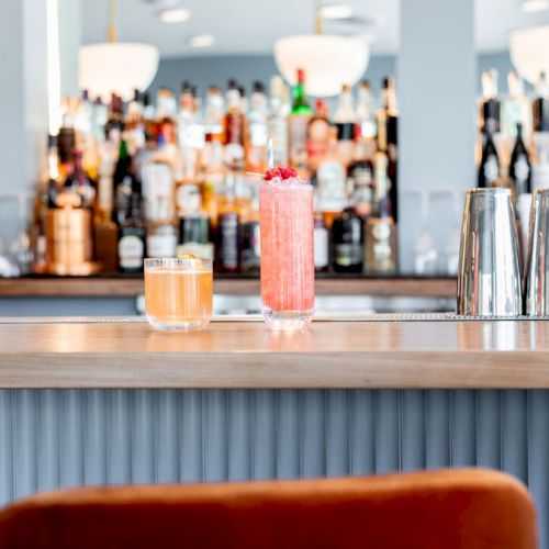 A bar counter with two cocktails, metal shakers, and shelves stocked with various bottles in the background, and a red chair in the foreground.