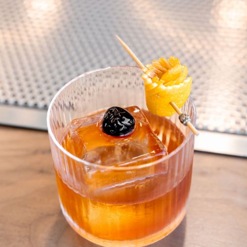 A cocktail in a glass with a large ice cube, garnished with a lemon twist and a cherry on a toothpick, placed on a bar counter.