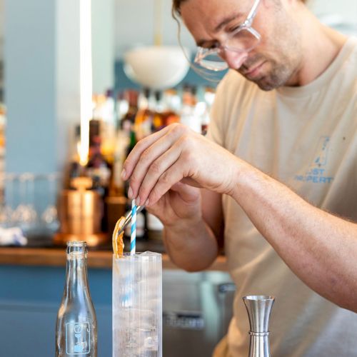 A person is meticulously preparing a drink behind a bar, using a bottle and various tools in a well-lit, modern setting.