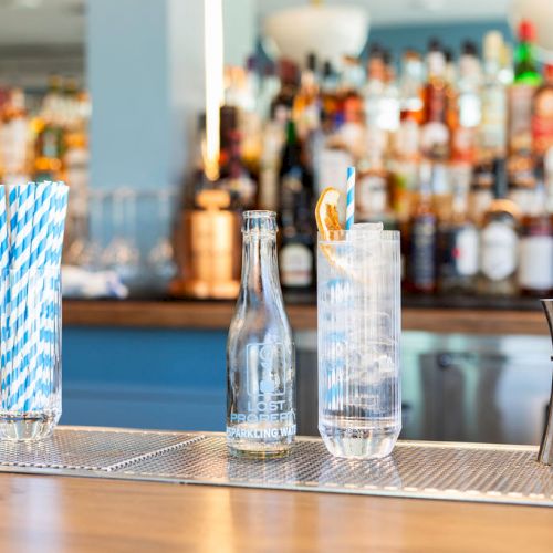 A bar counter with glasses, a bottle, a shaker, and straws. The background shows a variety of liquor bottles on shelves.