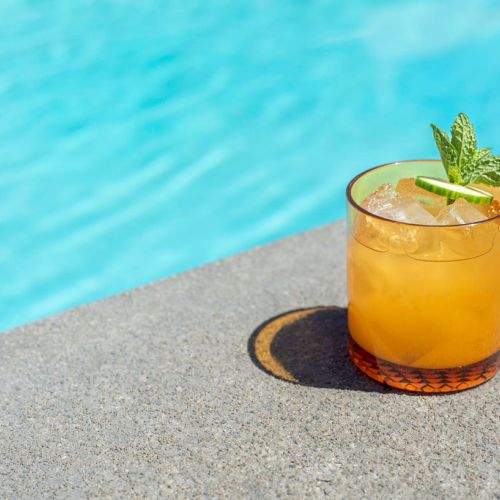 A refreshing orange-hued cocktail in a glass with ice, garnished with a lime slice and mint, placed next to a swimming pool.