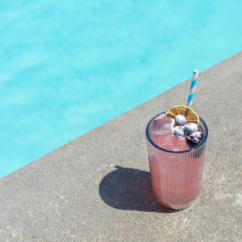 The image shows a pink drink with ice, garnished with fruit and a striped straw, placed on a poolside surface next to clear blue water.