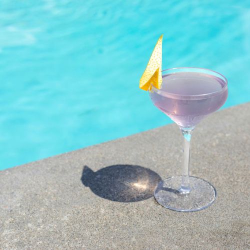 A purple cocktail with a lemon garnish sits on a concrete ledge next to a swimming pool, casting a shadow.