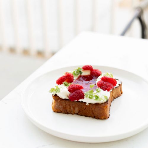 The image shows a slice of toast topped with whipped cream, raspberry jam, fresh raspberries, and a green garnish, served on a white plate.