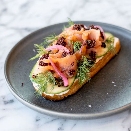 The image features an open-faced sandwich on a dark plate, topped with smoked salmon, herbs, pickled onions, and possibly caviar, on a marble surface.