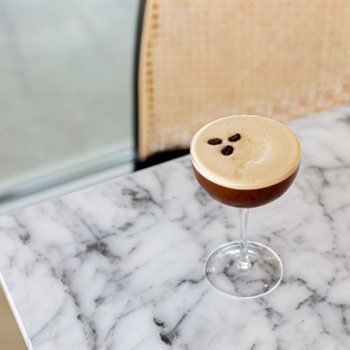 A cocktail glass with a frothy brown beverage topped with three beans, placed on a marble table next to a chair with a woven backrest.