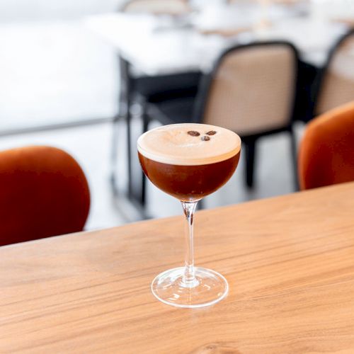 A cocktail in a stemmed glass with a frothy top and coffee beans on the foam sits on a wooden table with orange chairs in the background.