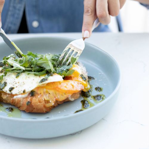 A person is cutting into an open-faced sandwich topped with eggs and greens, served on a gray plate, while seated at a table.