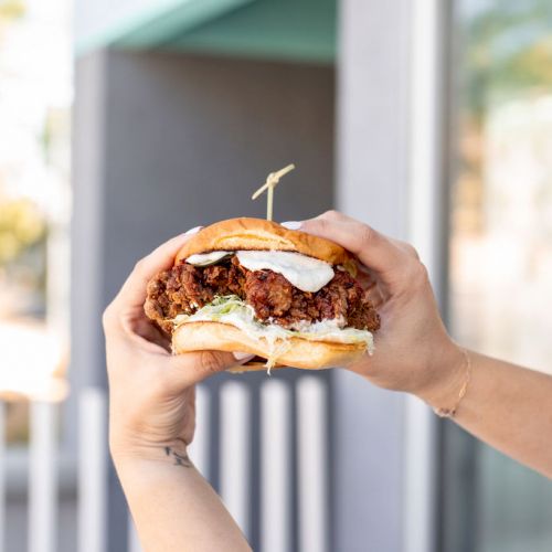 The image shows two hands holding a fried chicken sandwich with sauce and lettuce, secured with a toothpick, against a blurred outdoor background.