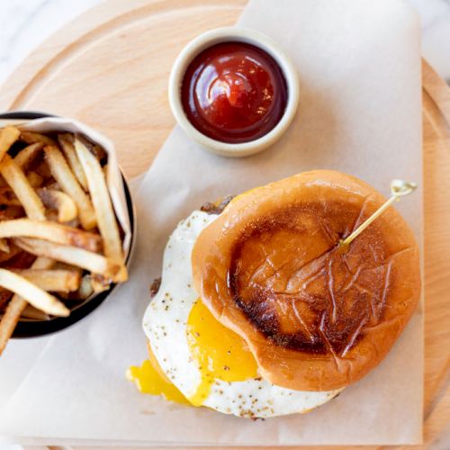 The image shows a burger with a fried egg, a side of fries in a container, and a small dish of ketchup on a wooden board.