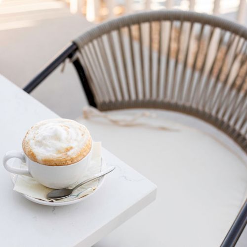 A cup of frothy cappuccino on a saucer with a spoon rests on a white table, next to an empty woven chair on a sunny day.