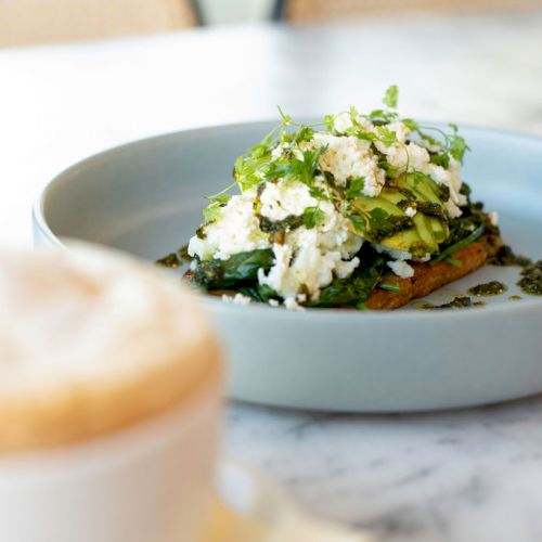 A plate with avocado toast topped with greens and cheese, and a cup of coffee on a marble table in the foreground.