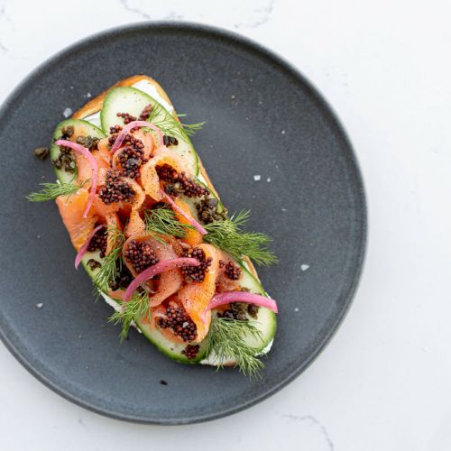 A dark plate holds an open sandwich with smoked salmon, dill, pickled onions, and presumably caviar, garnished with herbs.