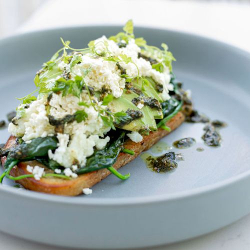 An open-faced sandwich with avocado, spinach, feta cheese, herbs, and a drizzle of sauce on a plate, garnished with fresh greens.