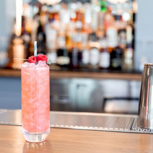 A tall pink cocktail garnished with berries is on the bar, with various bottles and shakers visible in the background.