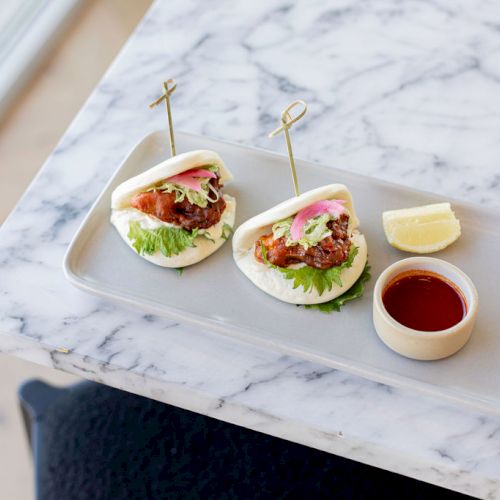 Two stuffed bao buns on a rectangular plate, garnished with a lemon wedge and a small cup of dipping sauce, are on a marble table.