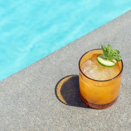 A refreshing cocktail garnished with a cucumber slice and mint sits on a poolside ledge next to a bright blue swimming pool.