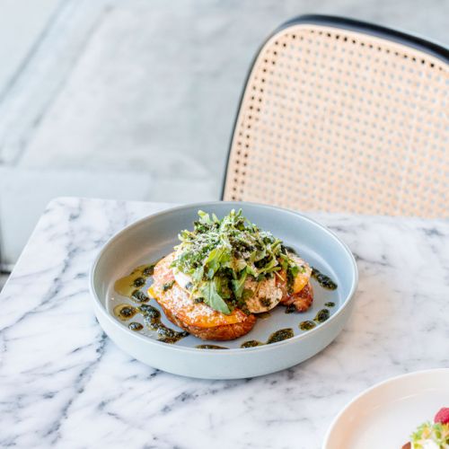 A plated gourmet dish topped with greens sits on a marble table next to a partially visible dessert plate and a woven chair.