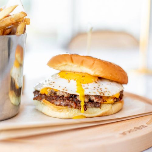 The image shows a cheeseburger topped with a fried egg on a wooden plate, accompanied by a container of French fries, with utensils to the side.