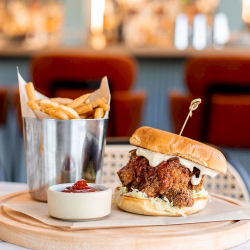 The image shows a fried chicken sandwich on a wooden board, accompanied by a metal bucket of fries and a small container of dipping sauce, in a restaurant.