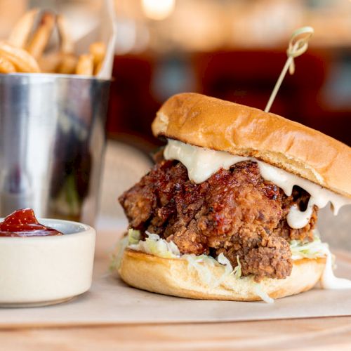 A fried chicken sandwich with lettuce and sauce, served with a side of fries in a metal cup and a small dish of ketchup.