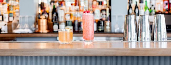 A bar's counter featuring two cocktails, several stainless steel shakers, and an array of bottles in the background.