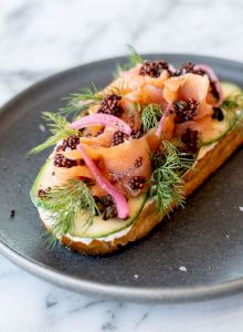 A piece of toast topped with smoked salmon, cream cheese, herbs, pickled onions, and caviar on a gray plate on a marble surface.