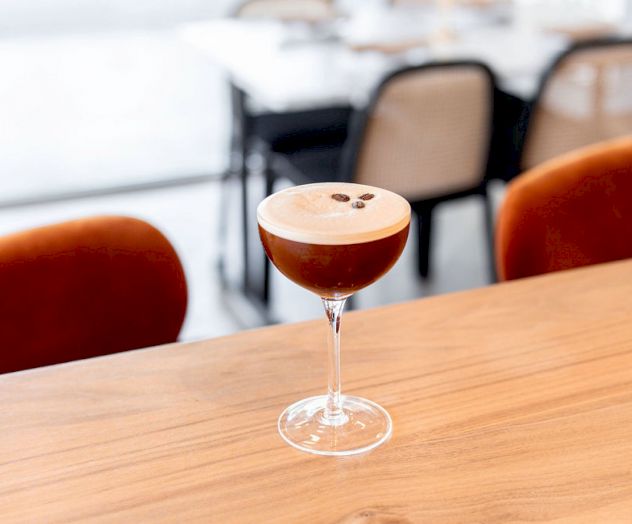 A cocktail with a creamy top, garnished with coffee beans, placed on a wooden surface in a modern, minimalistic setting.