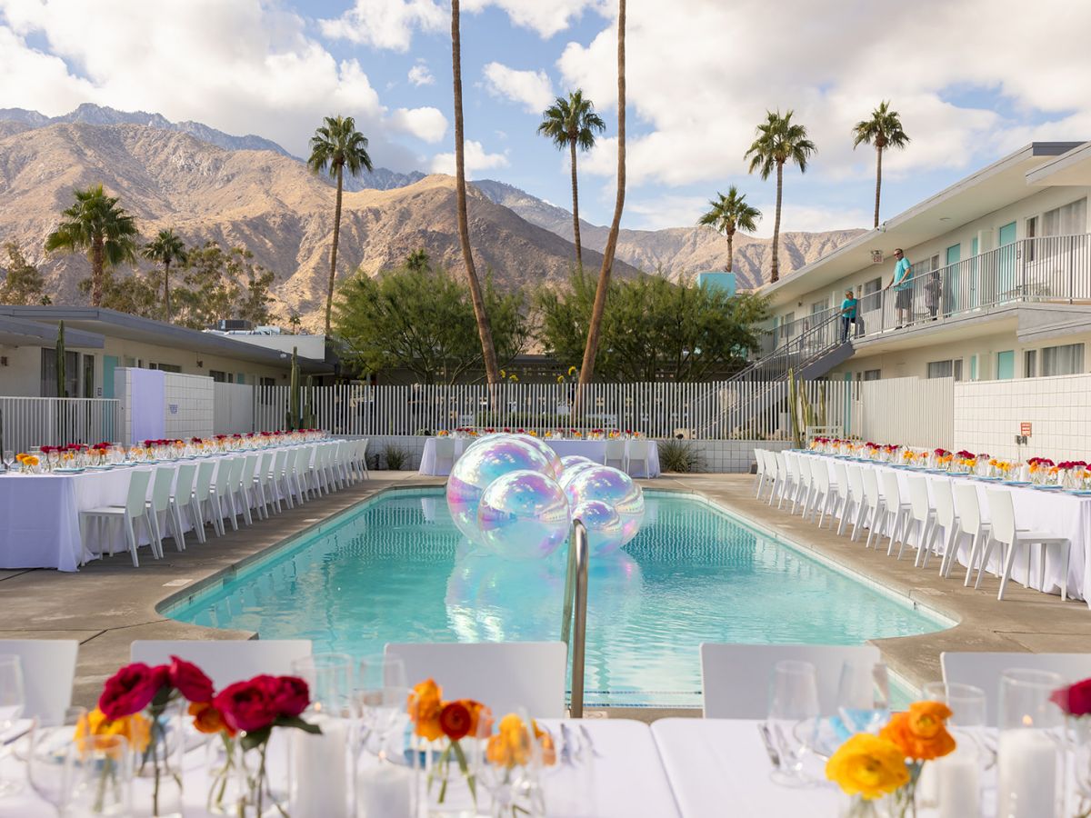 An outdoor setup with long tables next to a pool, decorated with colorful flowers and floating balloons, surrounded by palm trees and mountains.