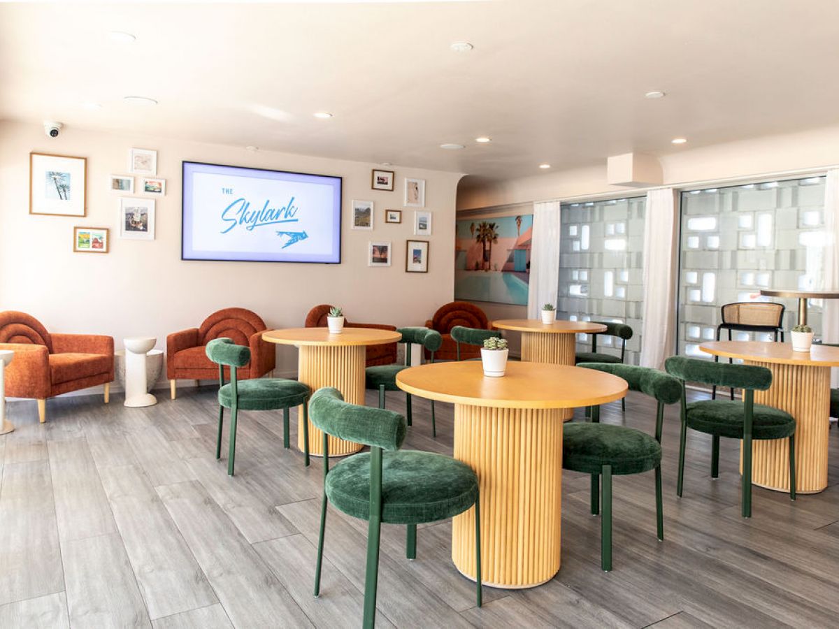 A modern, brightly lit cafe with round tables, green chairs, and wall art, featuring a screen displaying 