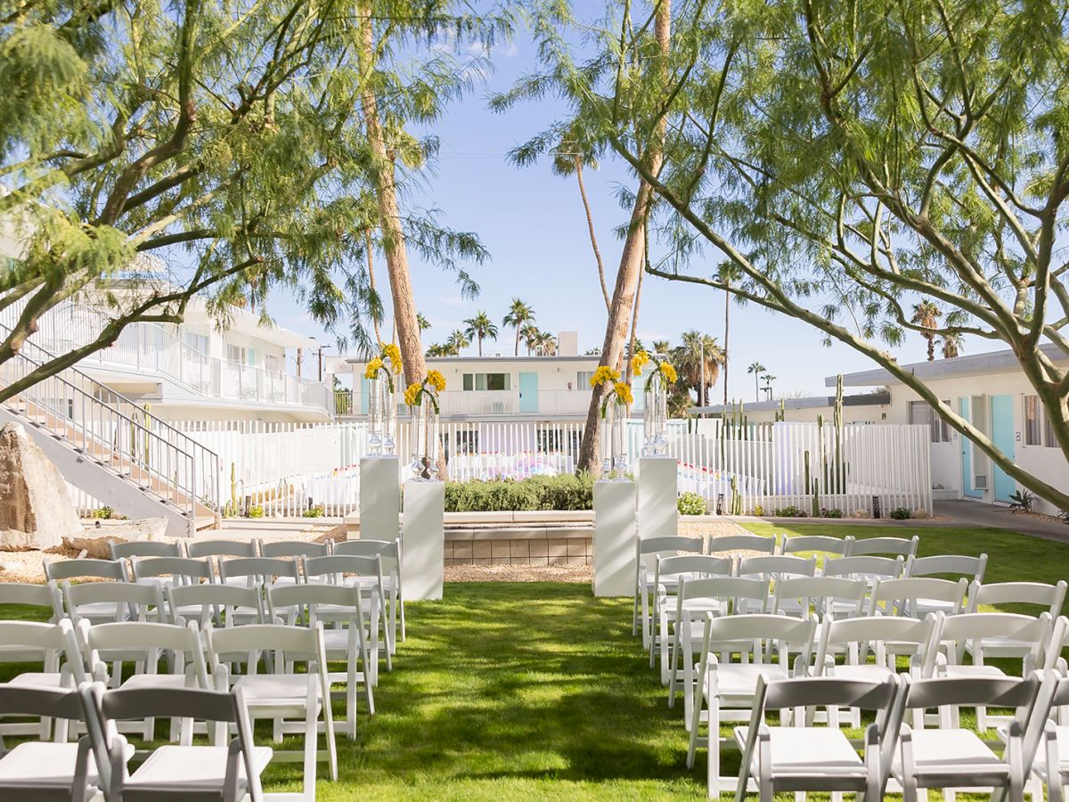 An outdoor wedding setup with white chairs organized neatly, facing an altar adorned with simple floral arrangements under trees.