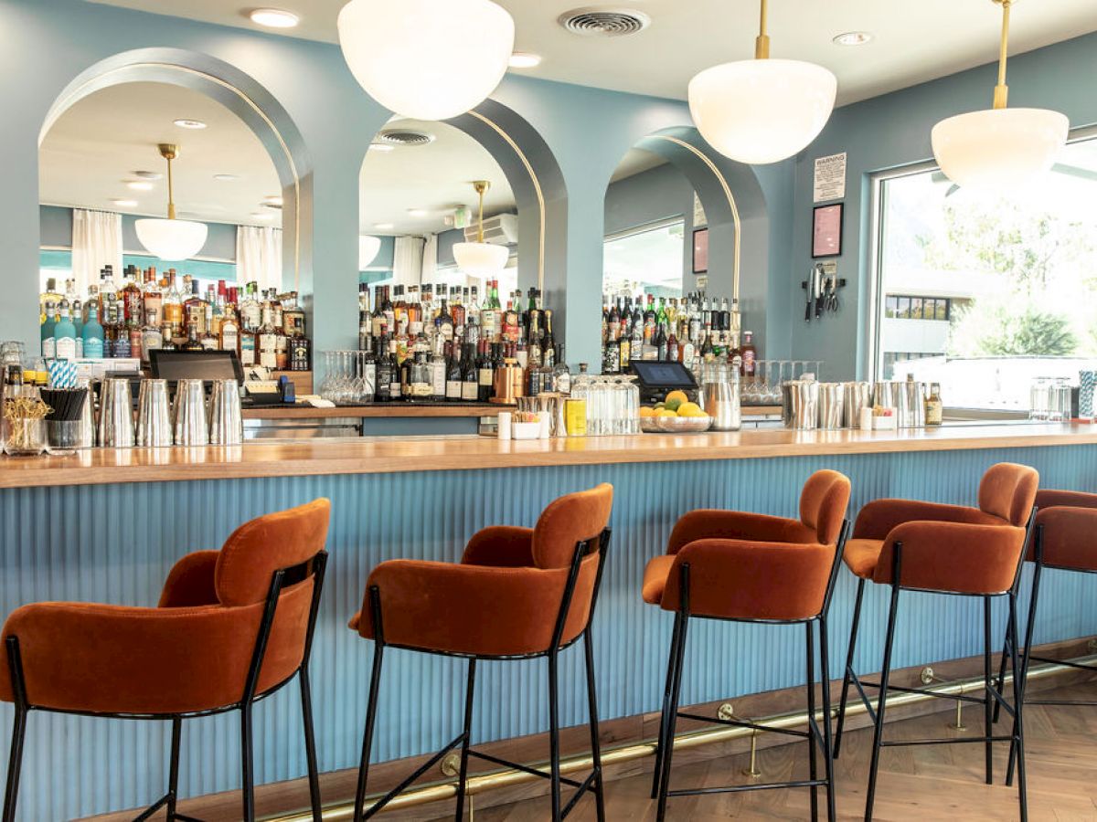 A stylish bar with a blue counter, orange chairs, and a well-stocked array of bottles and glasses. Bright pendant lights hang above.