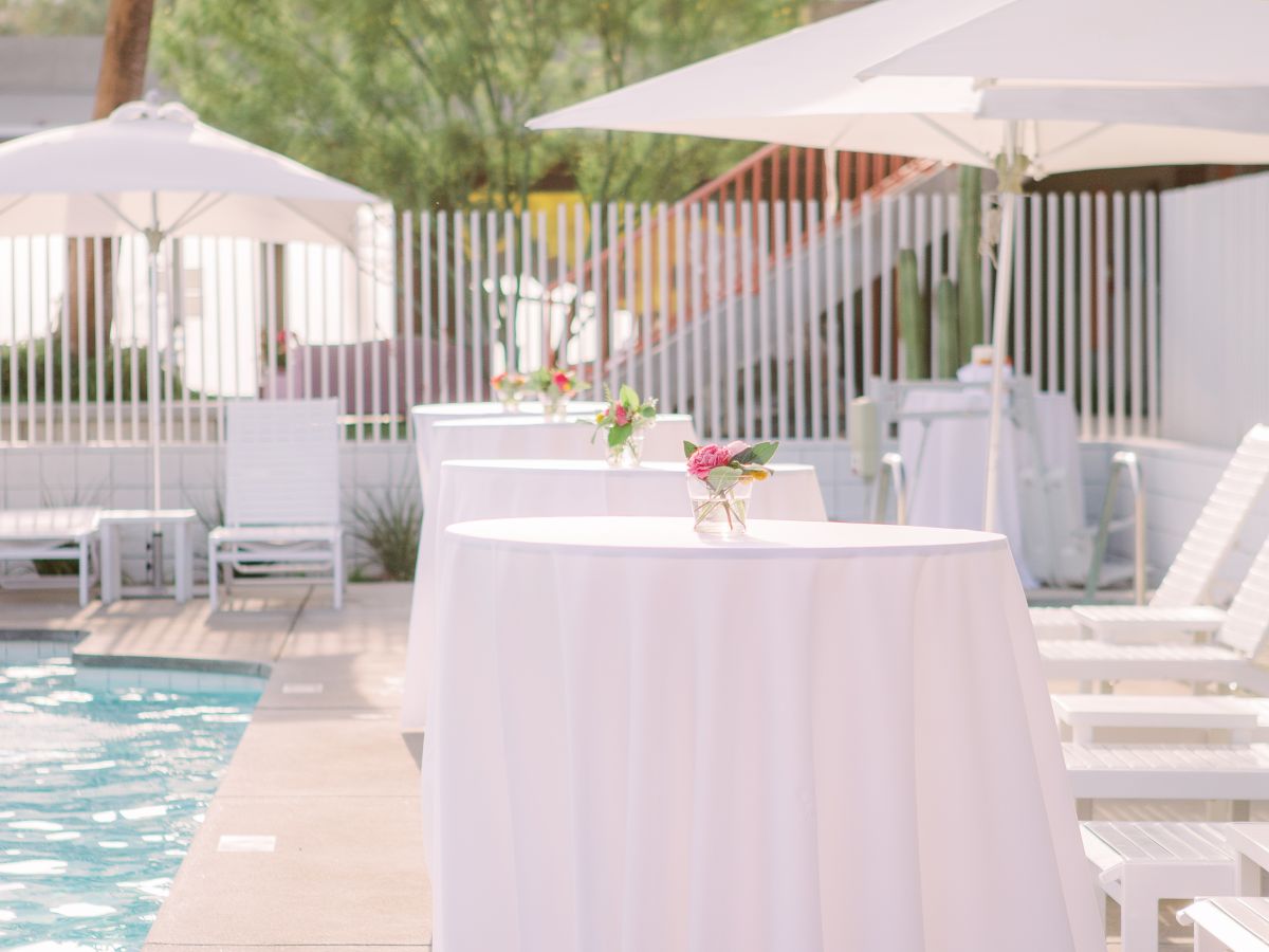 The image shows a poolside setting with white tables covered in cloth, lounge chairs, and umbrellas. There's a mountain in the background.