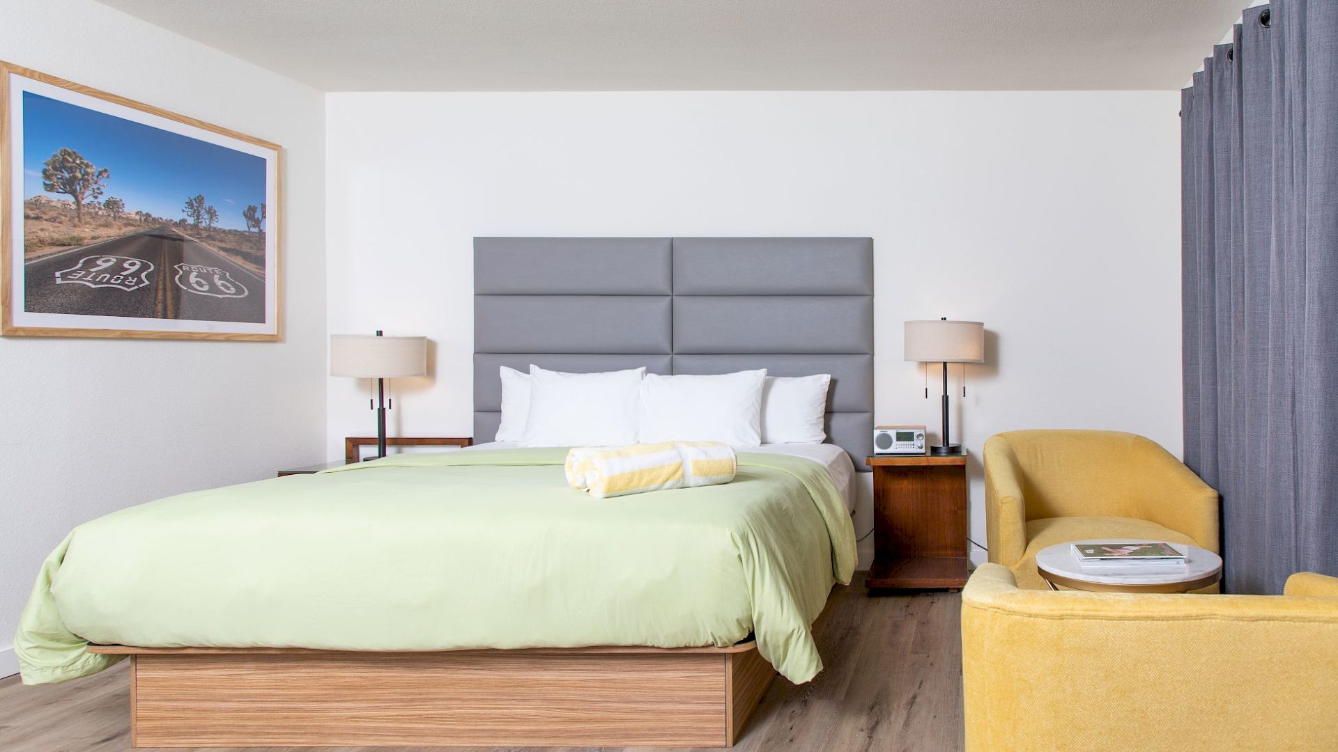 A modern bedroom with a green bed, two bedside tables, lamps, a yellow armchair, a landscape photo on the wall, and gray curtains.