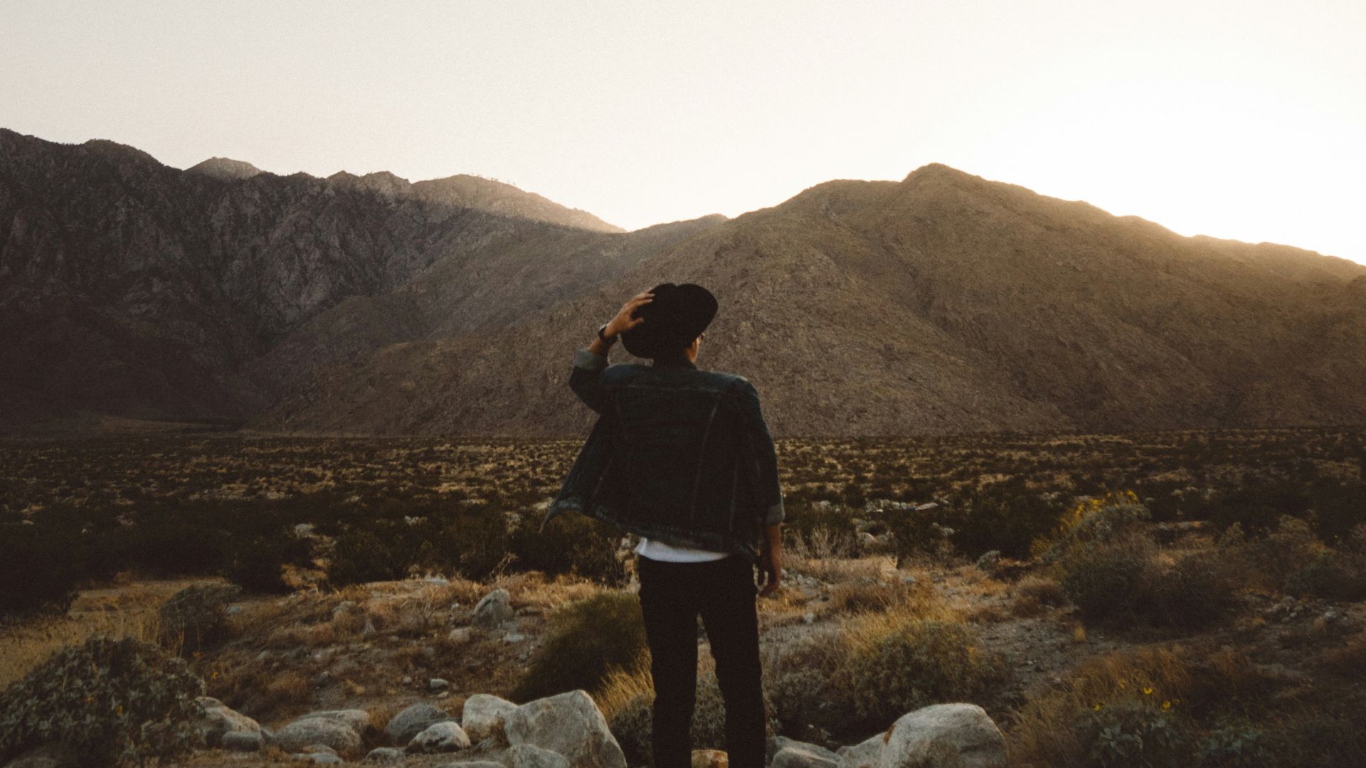 A person stands in a desert landscape with mountains in the background, captured during sunset or sunrise. They are facing away and wearing a hat.
