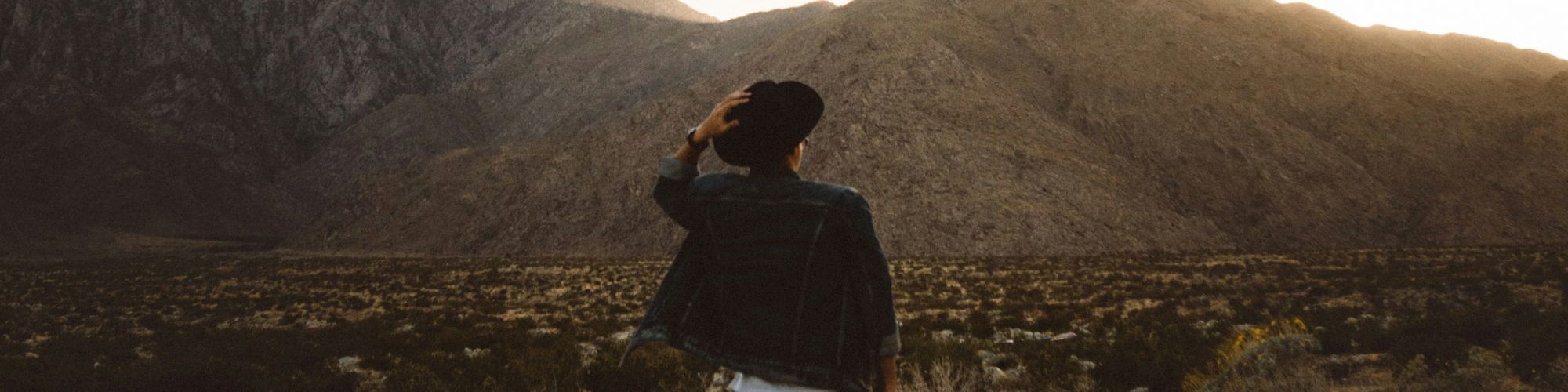 A person stands in a desert landscape with mountains in the background, captured during sunset or sunrise. They are facing away and wearing a hat.