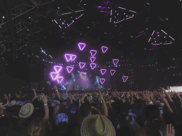 A large crowd enjoys a live concert with vibrant purple triangular lights on stage, creating a lively and energetic atmosphere.