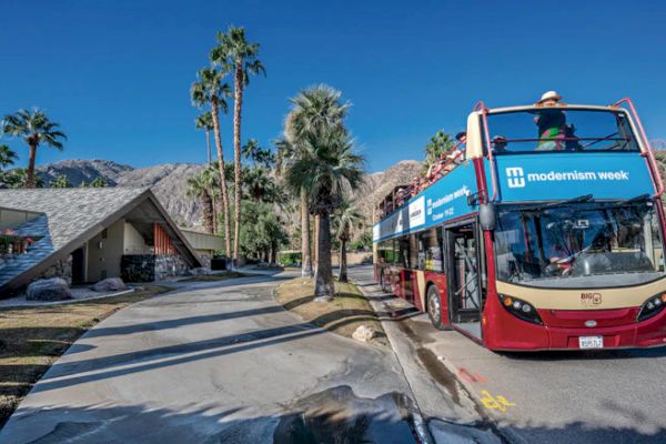 A double-decker bus labeled "Modernism Week" is parked on a sunny street near a modern-style house, with palm trees and mountains nearby.