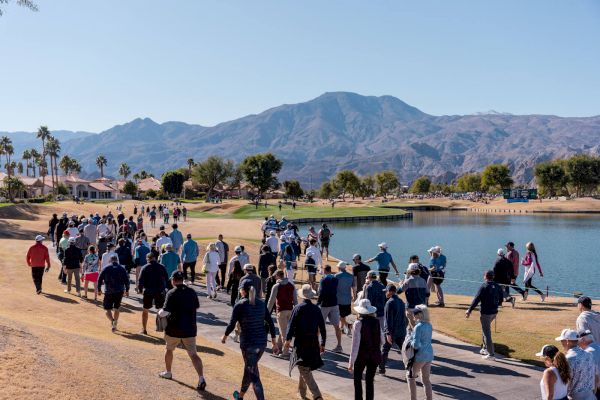 A large group of people walk along a path near a body of water with mountains in the background, under a clear blue sky.