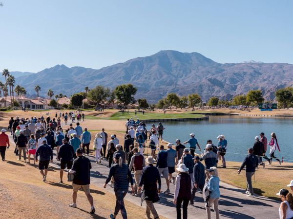 A large group of people walk along a path near a body of water with mountains in the background, under a clear blue sky.