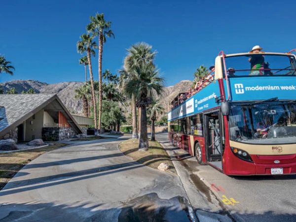 A double-decker bus labeled "modernism week" is parked on a palm-lined street, with mountains and a modern house in the background.