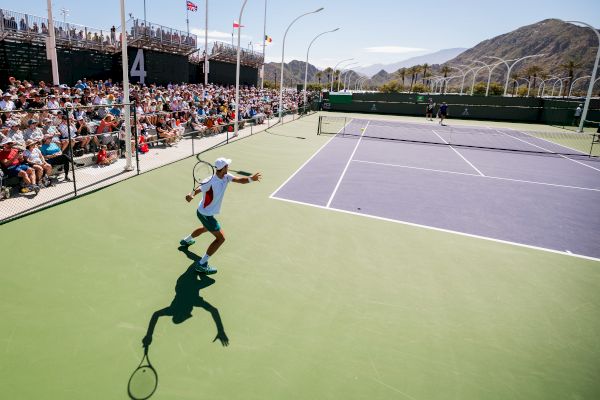 A tennis match is underway on an outdoor court with mountains in the background and a crowd watching from the stands.