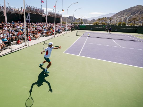 A tennis match is underway on an outdoor court with mountains in the background and a crowd watching from the stands.