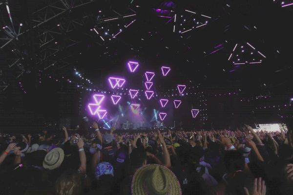 A crowd at a concert is gathered under a stage with purple geometric light displays, with hands raised towards the stage.