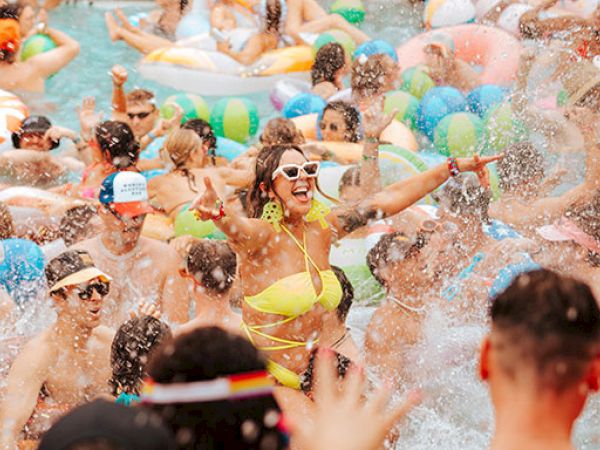 A crowded pool party with people swimming, splashing, and having fun. Inflatable toys and beach balls add to the lively atmosphere.