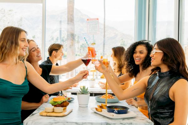 A group of friends toasts drinks at a restaurant table with food, enjoying a sunny day.